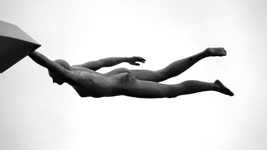 Black and white image of a sculpture depicting a figure diving, isolated against the sky.